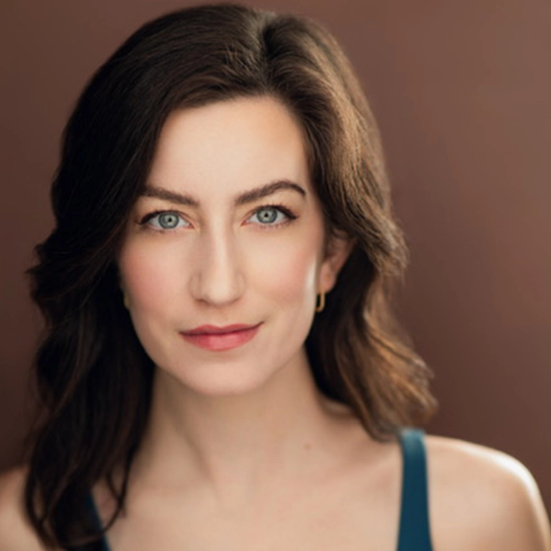 Kayleigh Brennan, a woman with wavy dark brown hair and blue eyes, looks directly at the camera. She has light skin, subtle makeup, and wears a blue sleeveless top. The warm brown gradient background creates a soft, professional portrait.