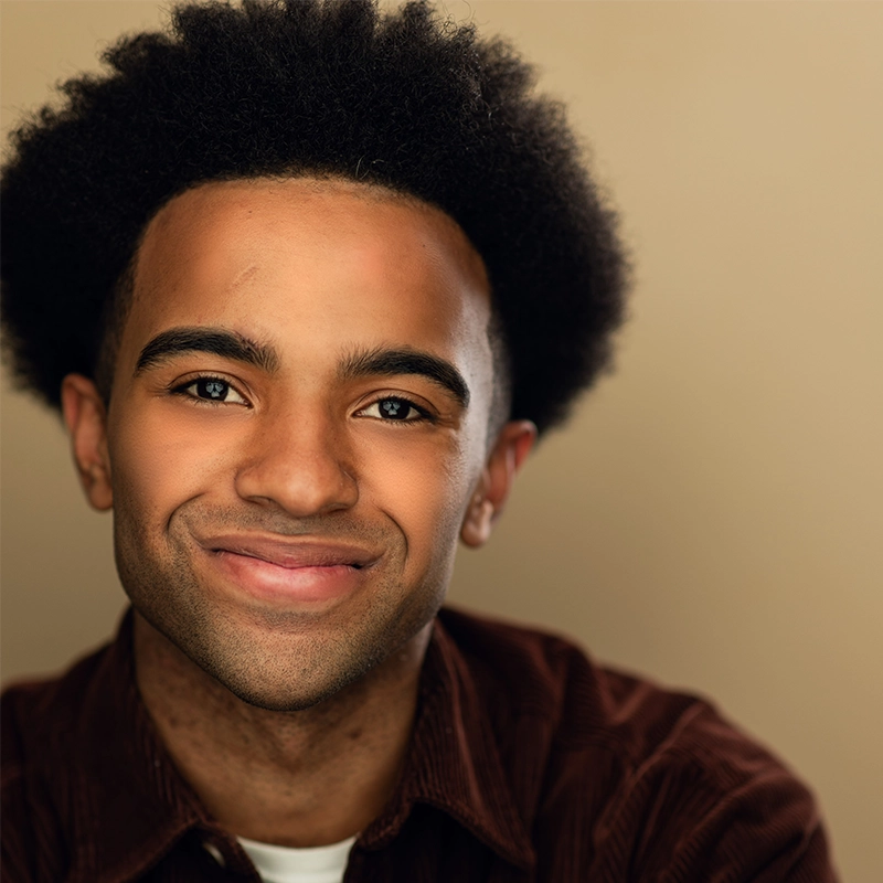 A young man with medium brown skin, short black curly hair, and a faint scar on his forehead smiles gently. He wears a brown corduroy shirt over a white T-shirt, and the background is a plain beige color.