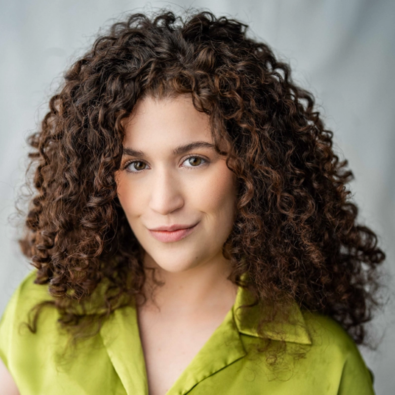 A woman with voluminous, curly brown hair smiles gently. She wears a lime green shirt with a collar. The background is softly blurred in neutral tones, drawing focus to her expressive eyes and natural makeup.