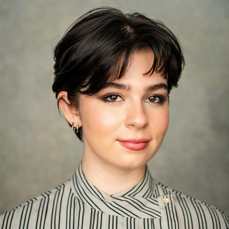 A young person with short dark hair and light skin smiles slightly. They wear subtle makeup, small hoop earrings, and a high-collared, cream-colored shirt with black vertical stripes. The background is softly blurred in neutral tones.