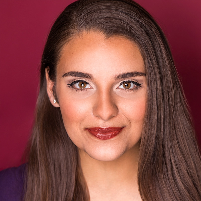 A woman with long, straight brown hair smiles softly. She wears bold eye makeup with eyeliner and dark red lipstick. She has brown eyes, stud earrings, and a maroon background behind her, creating a warm, polished portrait.