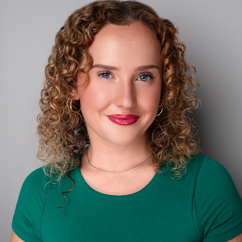 Molly Johnson, a woman with fair skin and curly, shoulder-length light brown hair, smiles gently. She wears a green top, gold hoop earrings, a delicate gold necklace, and natural makeup with red lipstick against a plain gray background.