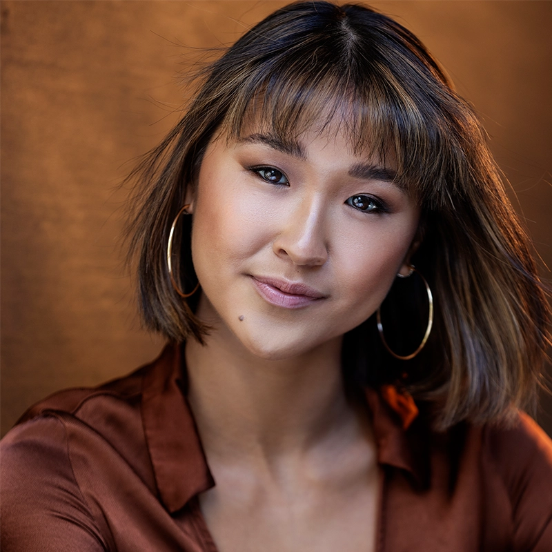 A young woman with straight brown hair and bangs, wearing large gold hoop earrings and a silky rust-colored top, faces the camera with a gentle smile against a softly blurred warm brown background.
