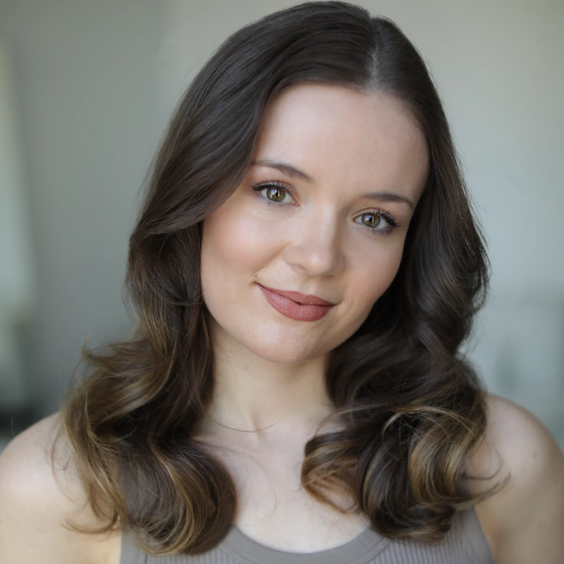 Lauren O'Brien, with wavy brown hair and fair skin, smiles softly at the camera. She wears natural makeup and a taupe tank top. The blurred background draws focus to her face and hair, which have subtle highlights.