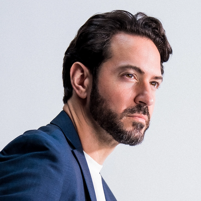 A man with short dark hair and a beard wears a navy blazer over a white shirt. Peter Douskalis looks thoughtfully to the right against a plain, light background.