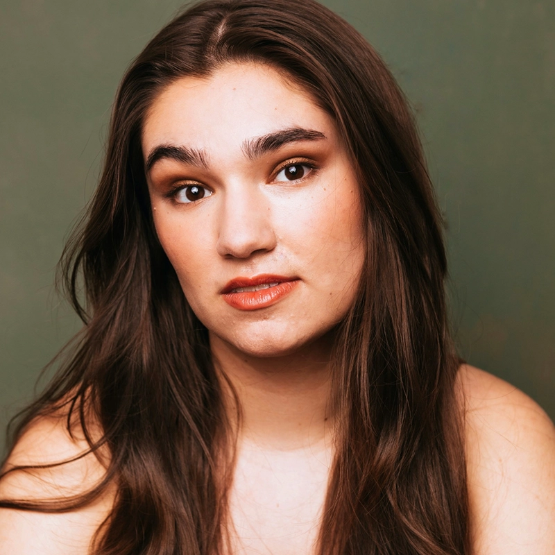A young woman with long brown hair and fair skin looks directly at the camera. She has natural makeup with brown eye shadow and red lipstick. The background is a soft green, and her expression is calm and neutral.