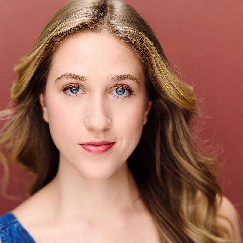 A young woman, Rachel Schoenecker, with long, wavy blonde hair and blue eyes looks directly at the camera. She has fair skin, light makeup, and wears a blue top. The background is a solid, muted reddish-brown. Her expression is calm and confident.