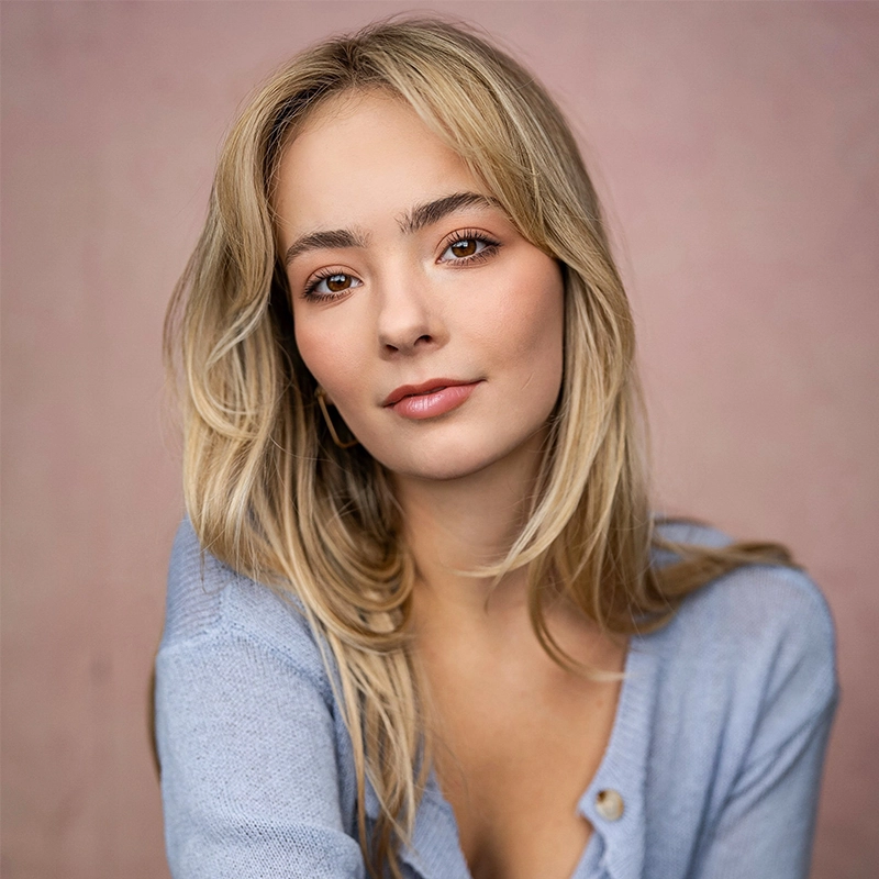 A young woman with long blonde hair and fair skin looks at the camera with a soft smile. She wears a light blue V-neck sweater and subtle makeup. The background is a muted pink, giving the image a gentle and warm atmosphere.