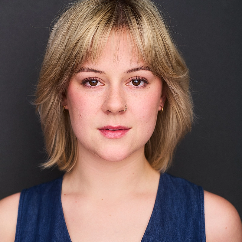 A young person with short, blonde hair and bangs faces the camera against a dark background. They have light skin, wear a nose ring, small hoop earrings, and a sleeveless blue top, with a neutral, calm expression.