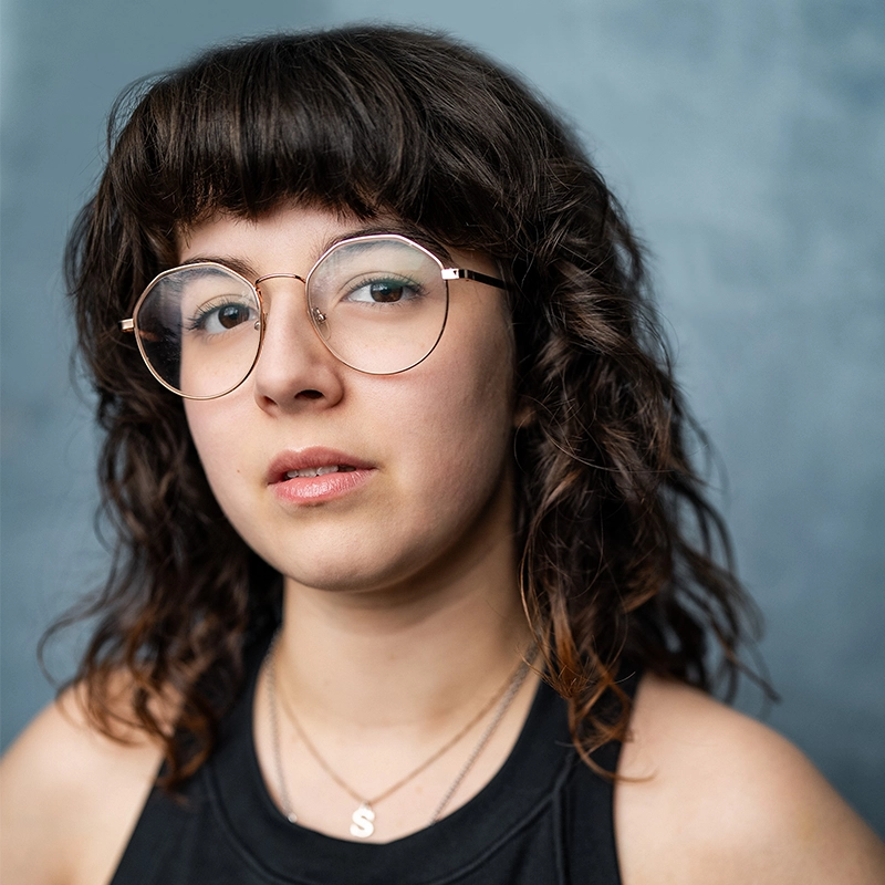A young person with wavy dark hair and bangs wears round glasses and a black sleeveless top. They have layered necklaces, one with an “S” pendant. The background is a smooth, light blue-gray gradient. They look confidently at the camera.