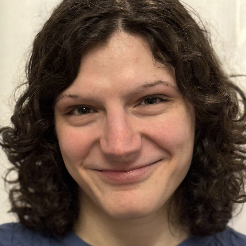 Matthew White, with short, curly brown hair, smiles softly at the camera. He has light skin, brown eyes, and wears a blue textured top. The softly blurred, light-colored background draws attention to his friendly expression.