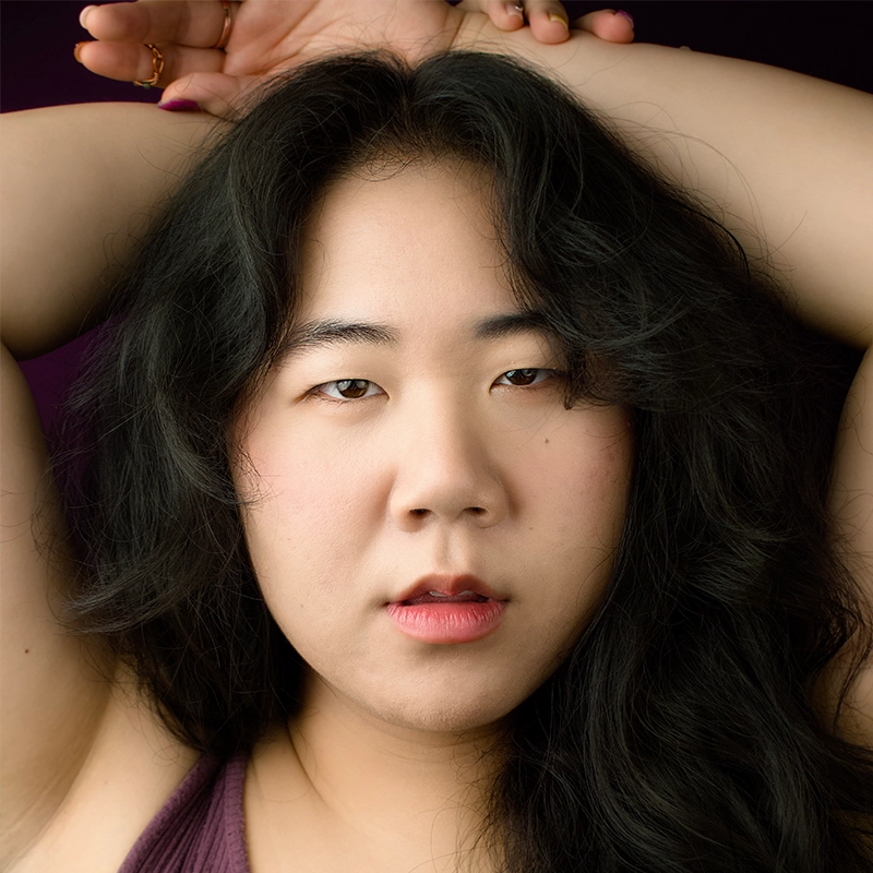 A person with long, wavy black hair poses with both arms raised above their head. They have light skin, subtle pink makeup, and parted lips. The background is dark, and they wear a sleeveless, mauve top. Their expression is calm and direct.