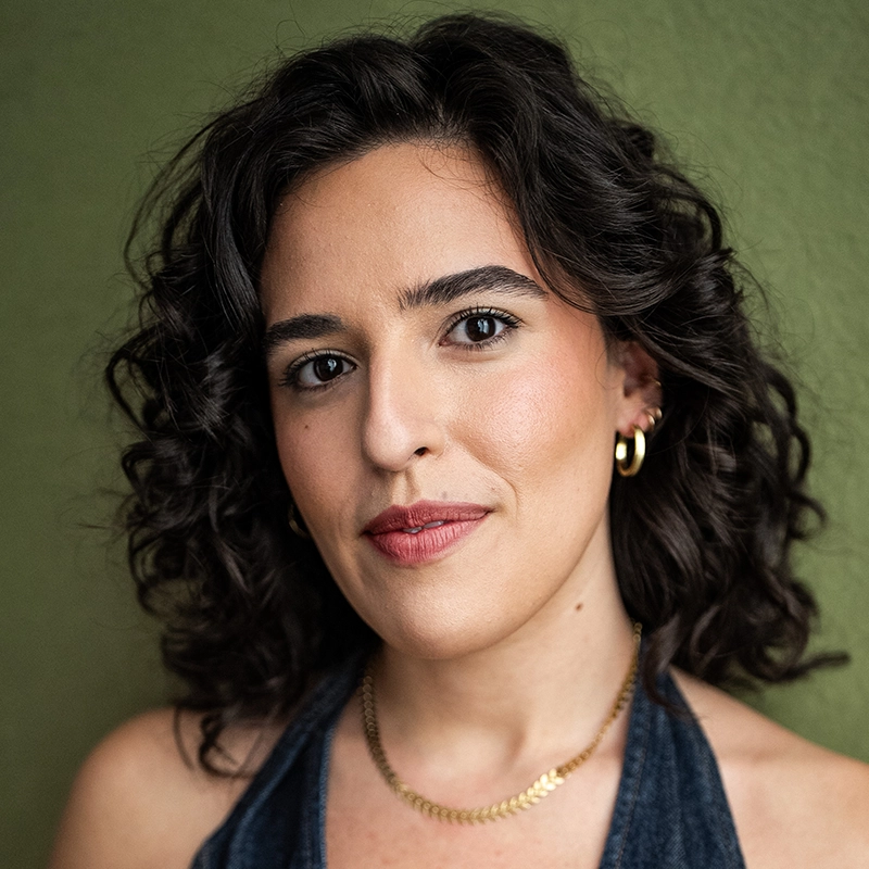 A woman with wavy dark hair and natural makeup smiles softly at the camera. Eleni Kontzamanys wears gold hoop earrings, a gold chain necklace, and a denim halter top, set against a plain olive green wall.