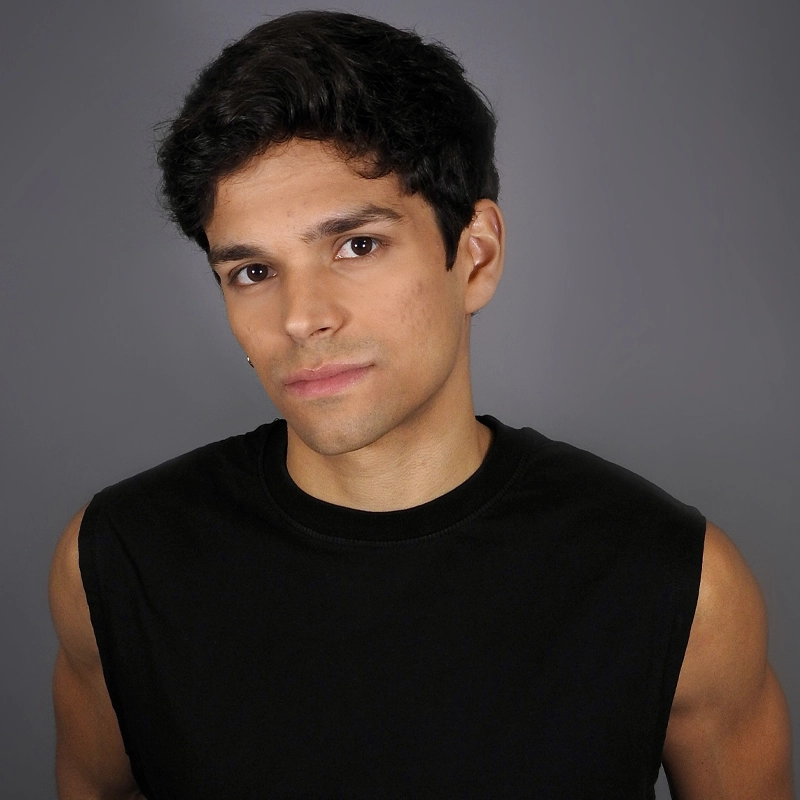 Miguel Amell, a young man with short dark hair and medium skin tone, stands against a plain gray background. He wears a sleeveless black shirt and looks at the camera with a neutral expression, slightly tilting his head to one side.