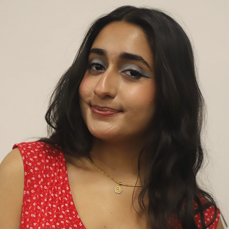 Anoushka Sharma, with long dark hair, wearing a red floral dress, a gold necklace, and blue eyeshadow, smiles subtly. She stands against a plain light-colored background, looking confidently at the camera.