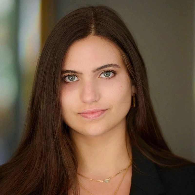 Kiana Mottahedan, a young woman with long brown hair and blue-green eyes, looks directly at the camera. She wears a black top, gold hoop earrings, and layered gold necklaces. The softly blurred background draws focus to her face.