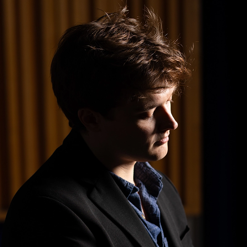 A young man with short, tousled brown hair is shown in profile against a dark background with vertical wooden slats—a portrait styled by Kiana Mottahedan. He wears a dark blazer over a blue shirt, softly lit from the side with gentle shadows on his face.