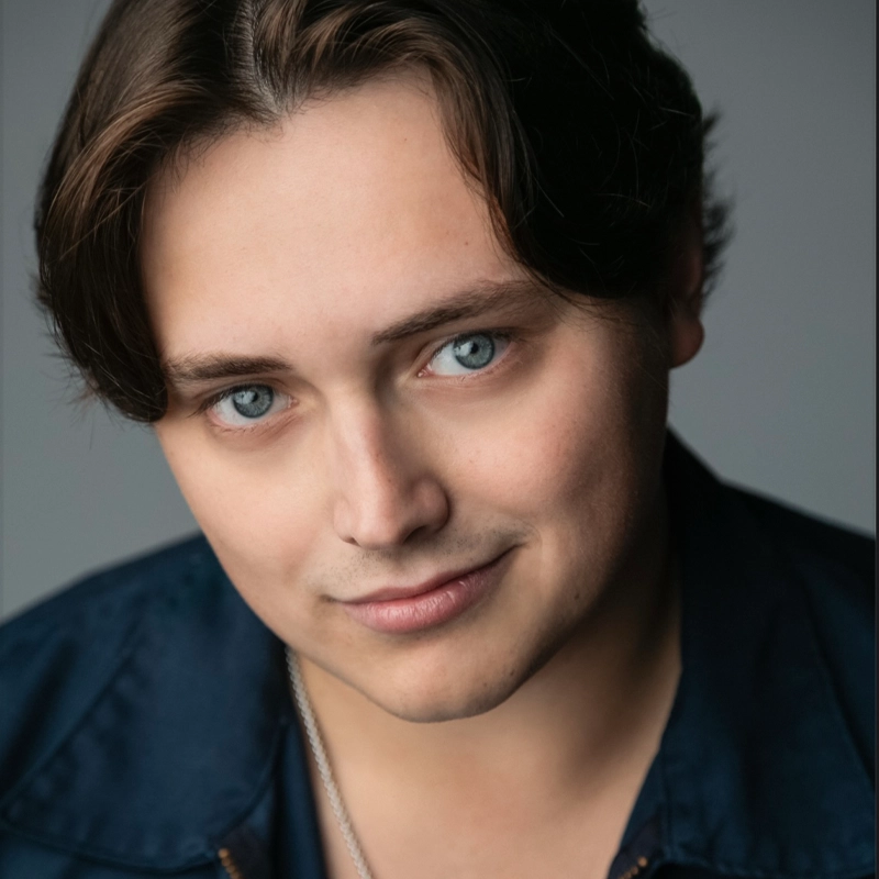A young person with fair skin, medium-length brown hair, and blue eyes, looking directly at the camera with a slight smile. Wearing a dark blue shirt and a thin necklace, Brandon Berg is set against a neutral gray background.