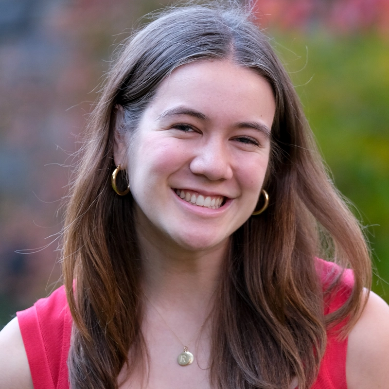 A young woman with long brown hair smiles outdoors. Rose Buchdahl wears gold hoop earrings, a gold necklace with a round pendant, and a sleeveless red top. The background is blurred with green and hints of red foliage.