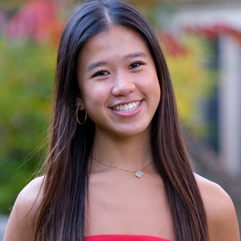 A young woman, Richelle Chang, with long straight brown hair smiles outdoors. She wears a red strapless top, gold hoop earrings, and a delicate gold necklace with a four-leaf clover pendant. The blurred background shows green and red foliage.
