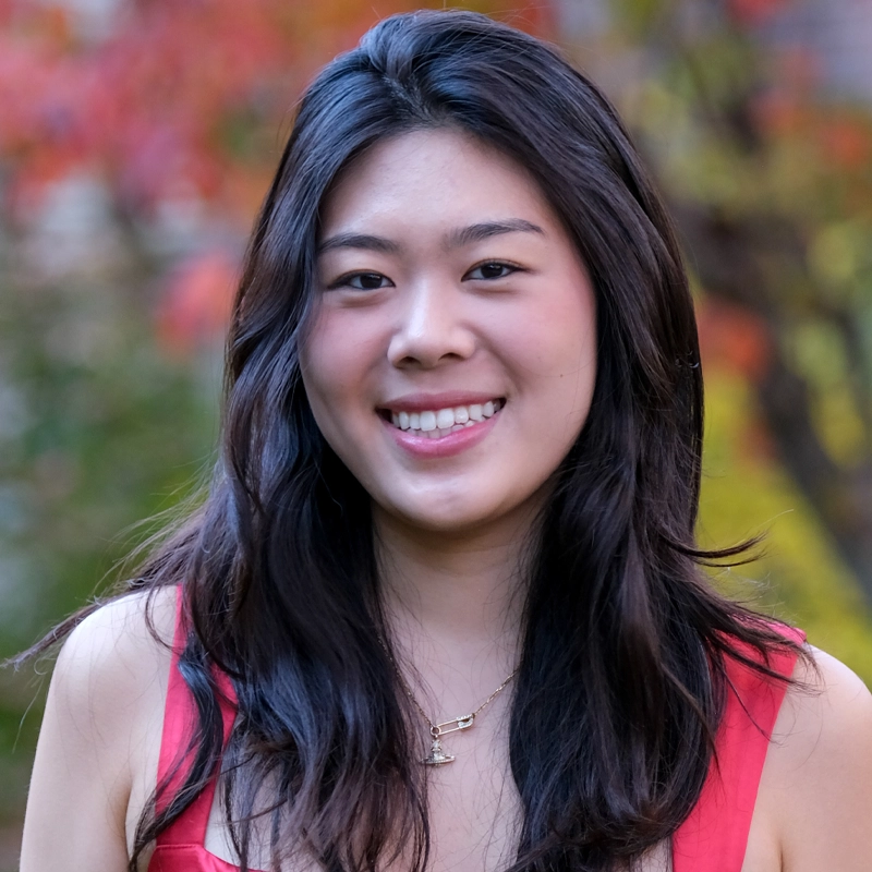 Michelle Cheon, a young woman with long, dark hair, smiles at the camera in a sleeveless red top and gold necklace. The blurred background reveals vibrant autumn foliage in shades of green, yellow, and red.