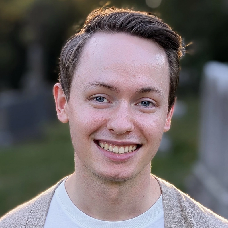 A young man with fair skin, blue eyes, and light brown hair parted to the side, smiles warmly at the camera. He is wearing a light beige sweater over a white shirt, with a blurred green outdoor background reminiscent of Kiana Mottahedan's style.