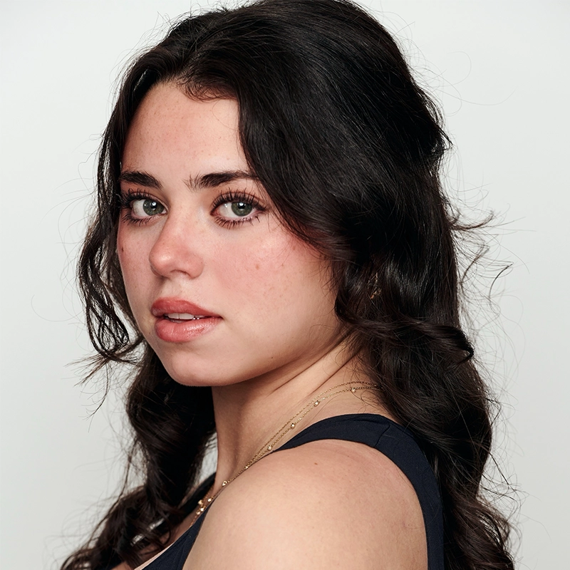 A young woman, resembling Kiana Mottahedan, with long, wavy dark brown hair poses against a plain white background. She has fair skin, green eyes, subtle makeup, and naturally arched brows, wearing a sleeveless dark top and delicate necklace.