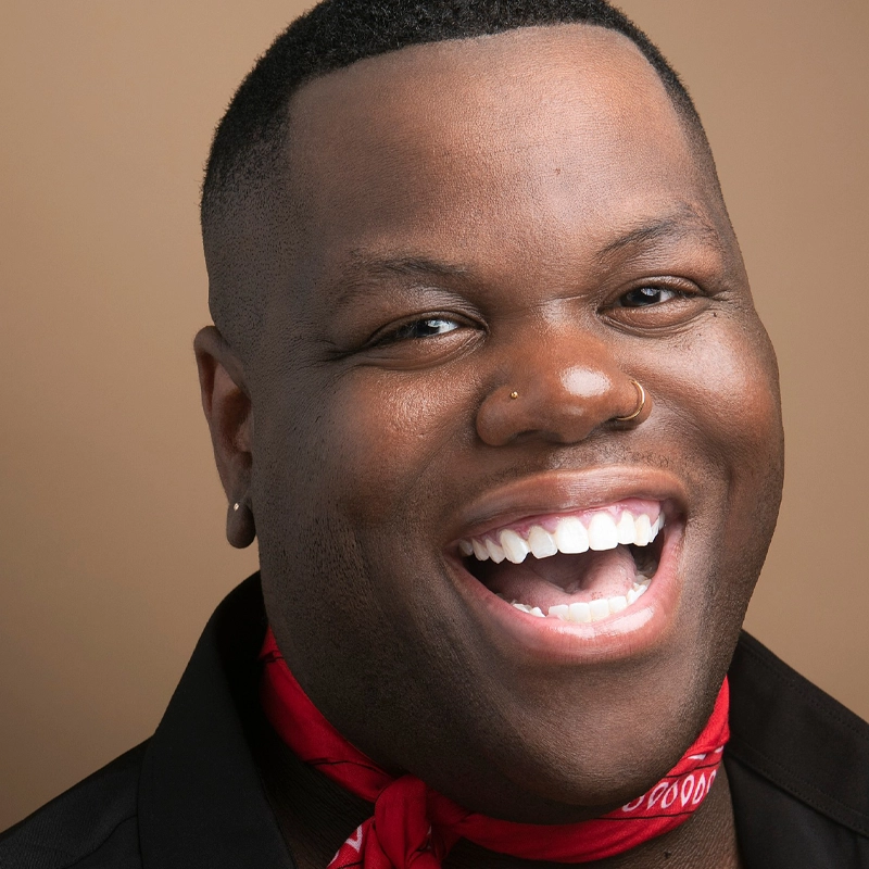 A person with dark skin and a close-cropped haircut smiles widely, showing their teeth. They wear a red bandana around their neck, a black top, a nose ring, and small earrings. The background is a plain, light brown color.