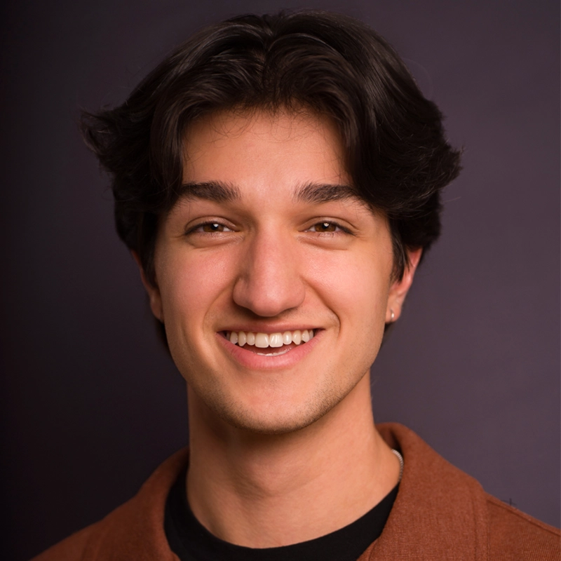 A young man with wavy dark hair smiles at the camera. He wears a brown shirt over a black top and a small hoop earring in his left ear. The plain, dark background creates a professional, studio-like portrait reminiscent of Kiana Mottahedan’s style.