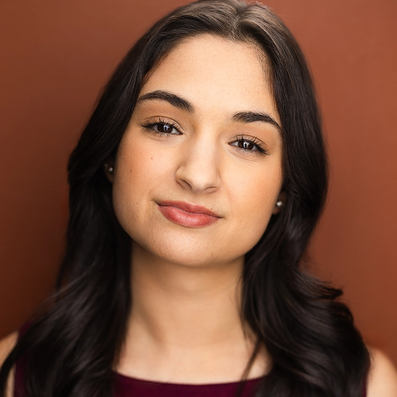 A young woman, Eleni Kontzamanys, with long, dark wavy hair and light skin is facing the camera against a warm brown background. She has subtle makeup, brown eyes, and a slight closed-mouth smile. She is wearing a sleeveless burgundy top.