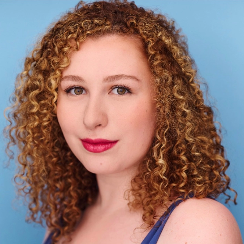 A woman with fair skin and shoulder-length, curly blonde hair is shown against a plain light blue background. She has brown eyes, subtle makeup, and wears a blue top with thin straps—Eleni Kontzamanys looks confidently at the camera.