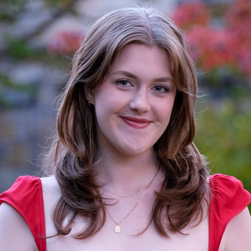A young woman with fair skin and wavy light brown hair smiles softly at the camera. She wears a red dress with cap sleeves and a delicate gold necklace. The blurred background features green foliage and hints of red leaves.