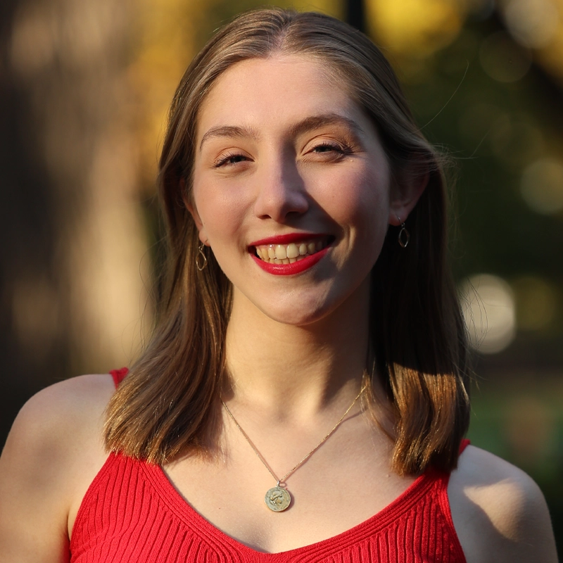 Lauren Fleissner, a young woman with straight, light brown hair, smiles outdoors in a red sleeveless top, gold hoop earrings, and a round gold pendant necklace. The blurred green and yellow background suggests a sunny day in the park.