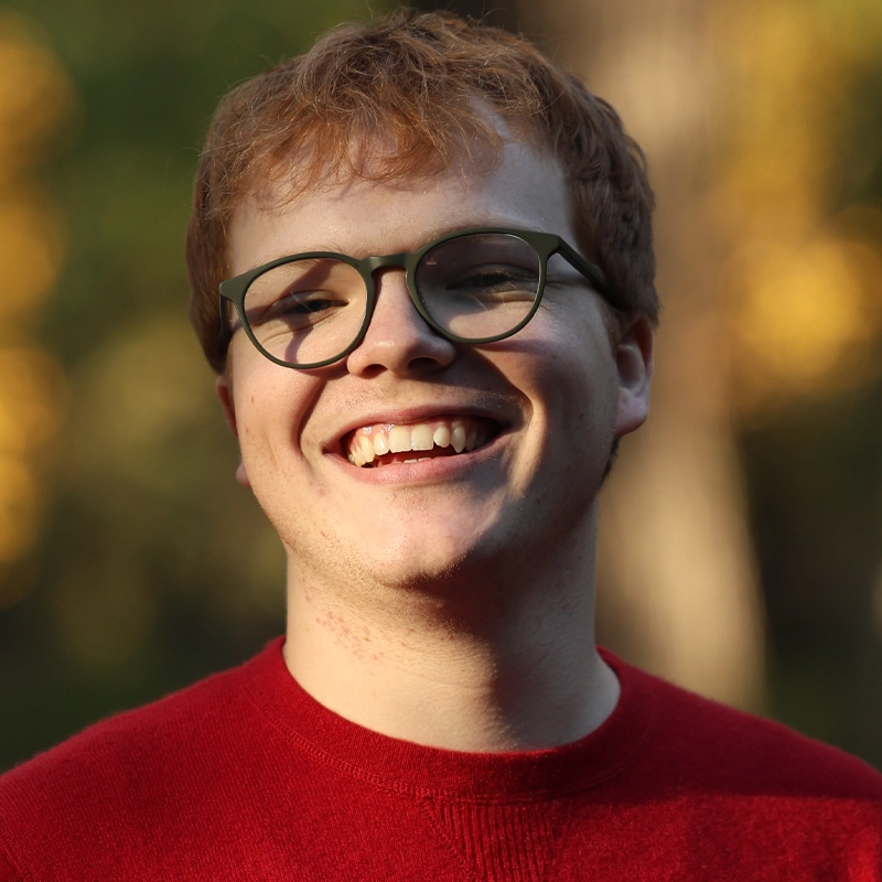 A young person with short, light red hair and glasses smiles broadly, showing teeth. They wear a bright red sweater and stand outside, with a blurred background of green and yellow, suggesting sunlight through trees.