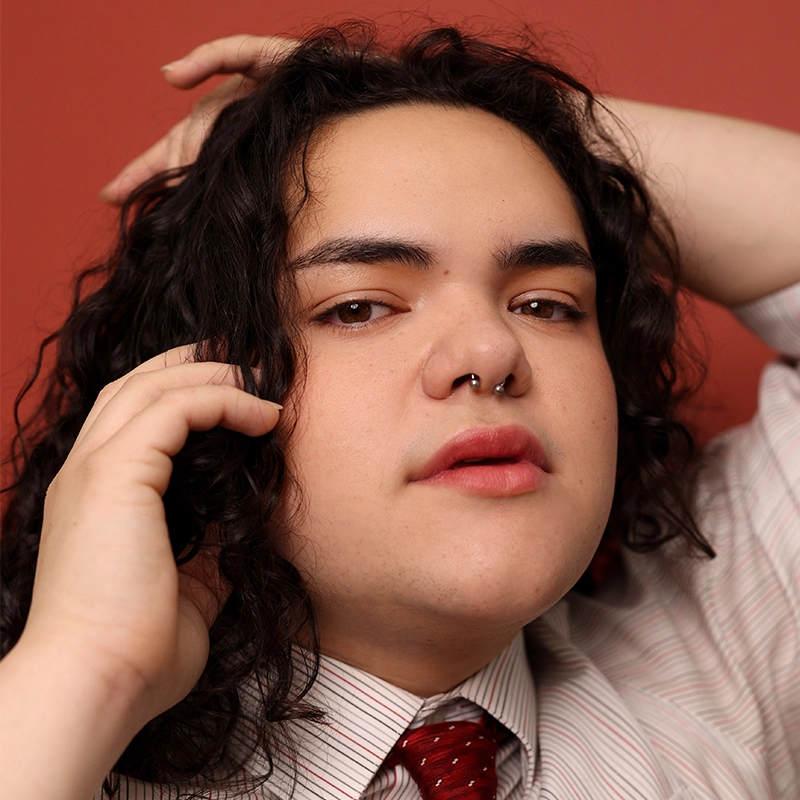 A person with long dark curly hair and a septum piercing poses with one hand on their head and the other touching their hair. Eleni Kontzamanys wears a white striped shirt and red patterned tie against a warm reddish background.