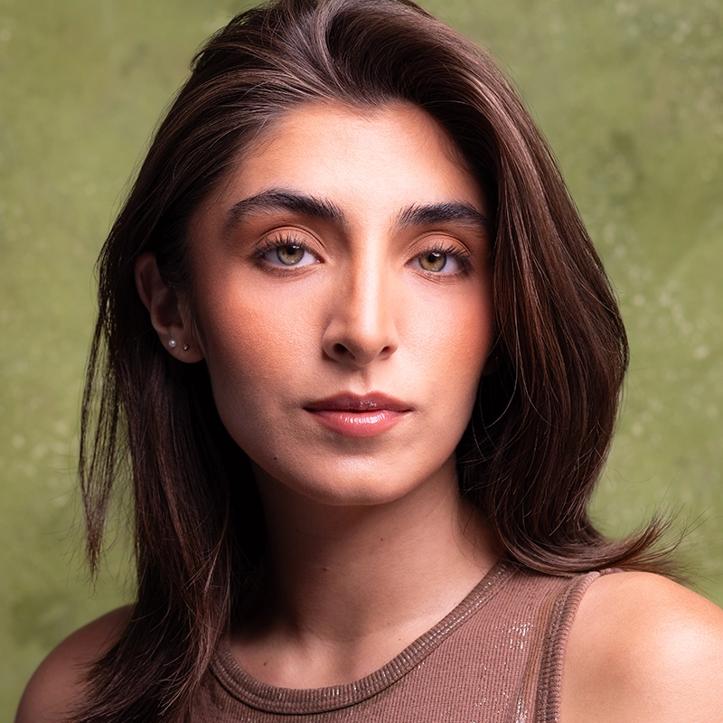 Kiana Mottahedan, a young woman with straight brown hair, light skin, and hazel eyes, looks at the camera with a neutral expression. She wears a sleeveless brown top and subtle makeup against a textured, muted green background.