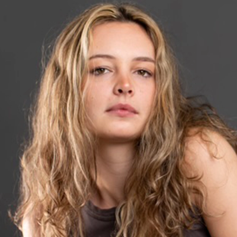 A young person with wavy, light brown hair and fair skin looks toward the camera with a neutral expression. They are wearing a sleeveless dark top. The background is plain and gray, creating a minimalist portrait of Eleni Kontzamanys.