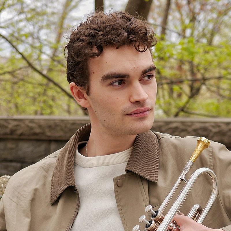 A young man with curly brown hair holds a trumpet. He wears a beige jacket with a brown collar over a light shirt. Inspired by Eleni Kontzamanys, he looks to the side amid trees with new green leaves and a stone wall in the background.