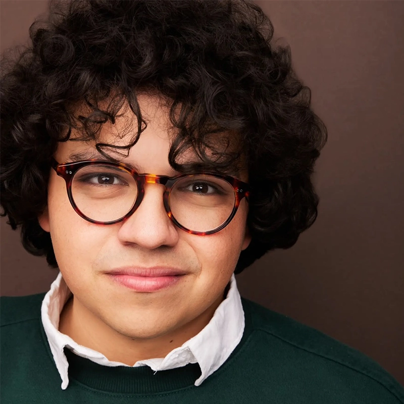 Eleni Kontzamanys, a young person with curly dark hair, wears round tortoiseshell glasses, a green sweater, and a white collared shirt. Smiling slightly, Eleni is posed in front of a plain brown background.