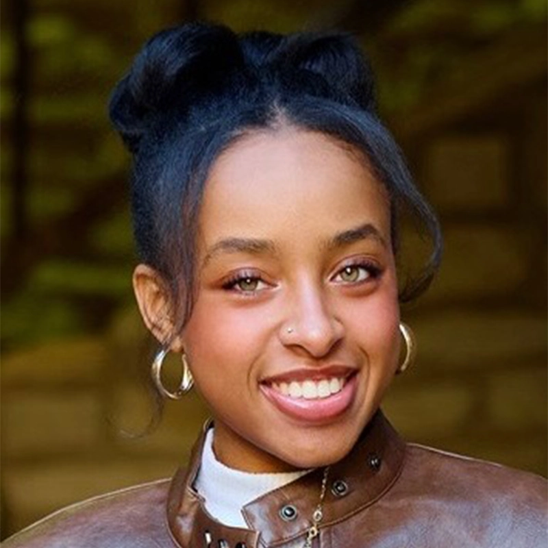 Kiana Mottahedan, a young woman with dark hair styled in two buns, smiles at the camera. She wears gold hoop earrings, a nose stud, a white turtleneck, and a brown leather jacket against an outdoor backdrop of green and brown tones.