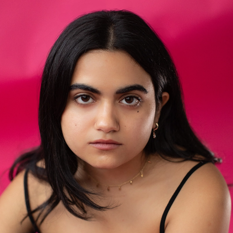 A young woman, Kiana Mottahedan, with straight dark hair and a neutral expression looks directly at the camera. She wears a thin-strapped black top, delicate necklace, and small earrings. The solid bright pink background adds a vibrant, modern feel.