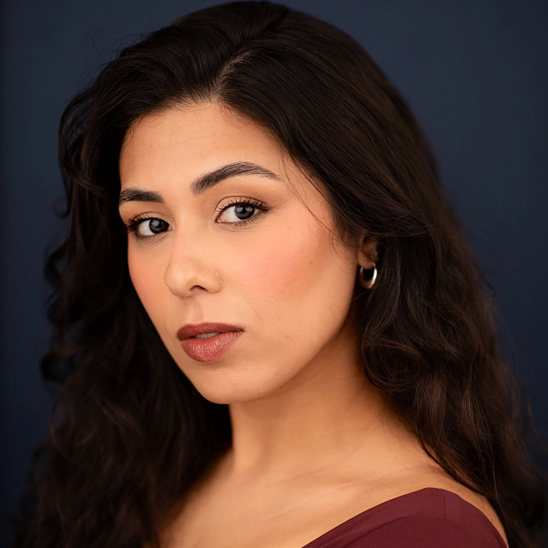 A woman with long, wavy dark hair poses against a dark blue background. Wearing a burgundy top and a small hoop earring, Eleni Kontzamanys gazes seriously at the camera, her natural makeup featuring defined brows and mauve lipstick.