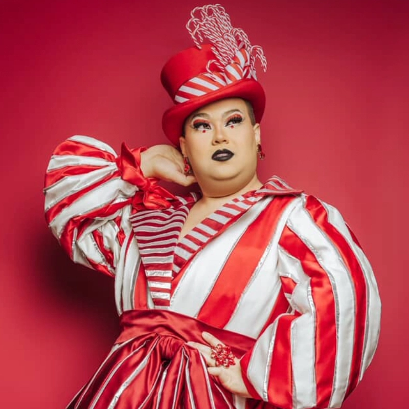 Lady Celestina poses confidently against a red background, wearing a dramatic red and white striped gown with puffed sleeves and a wide sash. She completes the look with a tall red hat adorned with white feathers, bold makeup, and striking black lipstick.