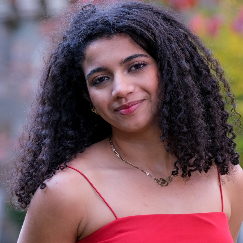 Sophia Lahik, with curly black hair, wearing a red spaghetti strap dress and a gold necklace, smiles softly at the camera. The background is blurred, with hints of greenery and colorful foliage.