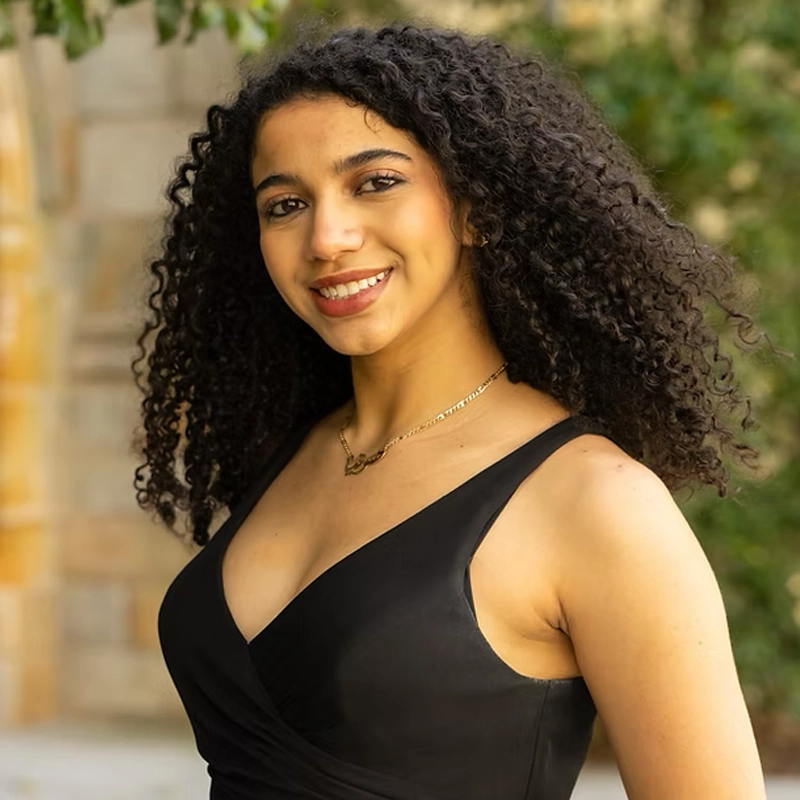 Sophia Lahik, a young woman with long, curly dark hair, smiles outdoors. She wears a sleeveless black dress and a gold necklace. The softly blurred background of greenery and a stone wall highlights her as the main focus of the image.