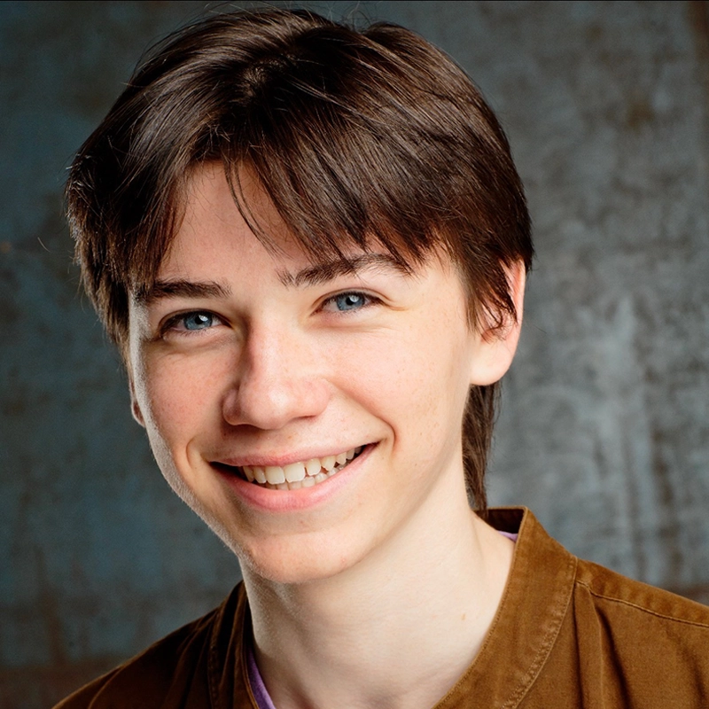 A smiling teenage boy, Luca Tuana, with straight brown hair and blue eyes, wearing a brown collared shirt over a light t-shirt, stands in front of a textured gray-blue background. The bright lighting highlights his cheerful expression.