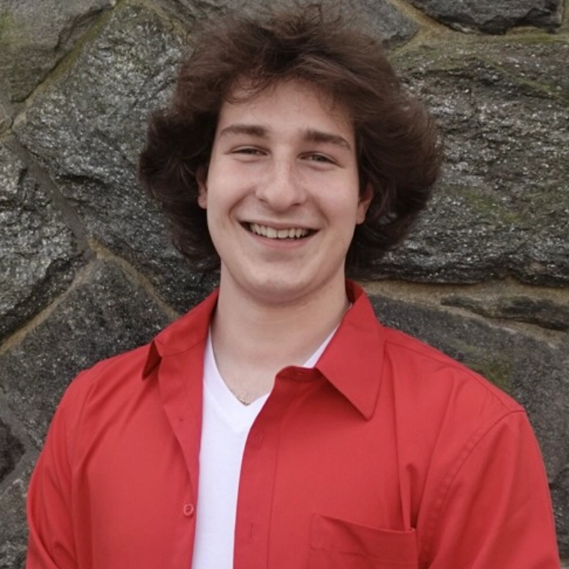A young man with wavy brown hair smiles at the camera. Brandon Macaluso is wearing a red button-up shirt over a white t-shirt, standing in front of a stone wall made of large, uneven rocks.