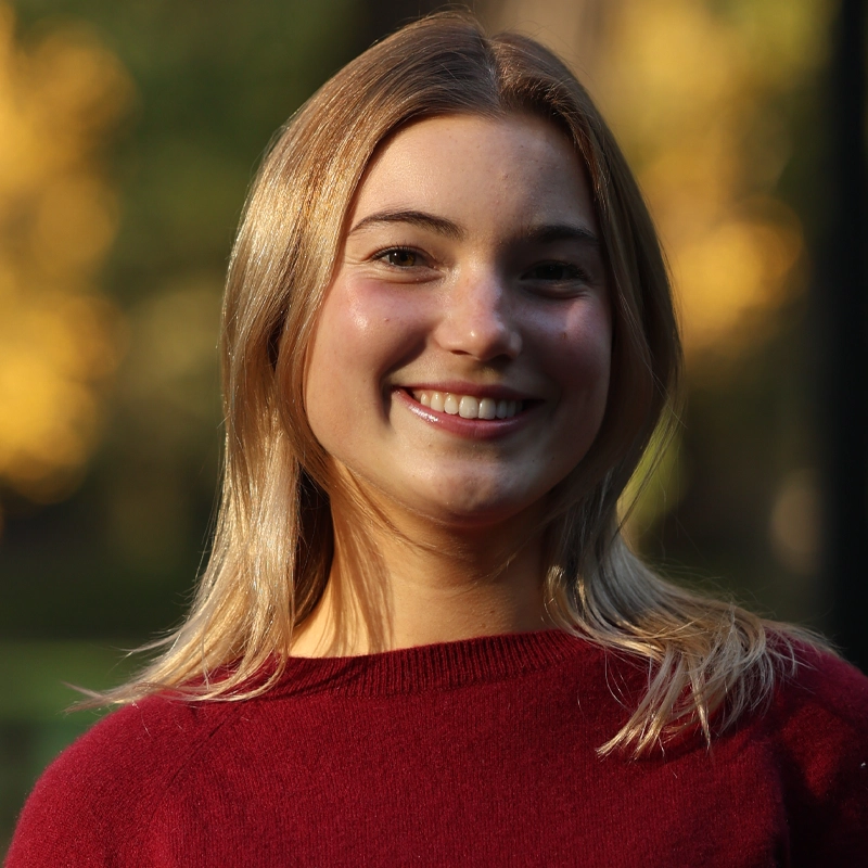 Estella Maguire, a young woman with straight blonde hair, smiles warmly at the camera. She is wearing a red sweater and is outdoors, surrounded by green trees and patches of yellow sunlight that highlight her face and hair.
