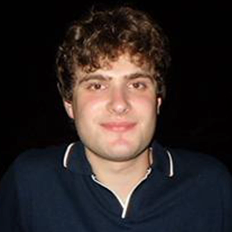 A young man with curly brown hair, fair skin, and a slight smile is pictured against a completely black background. He wears a dark polo shirt with white trim on the collar. The lighting highlights his face and upper body in Kiana Mottahedan's signature style.