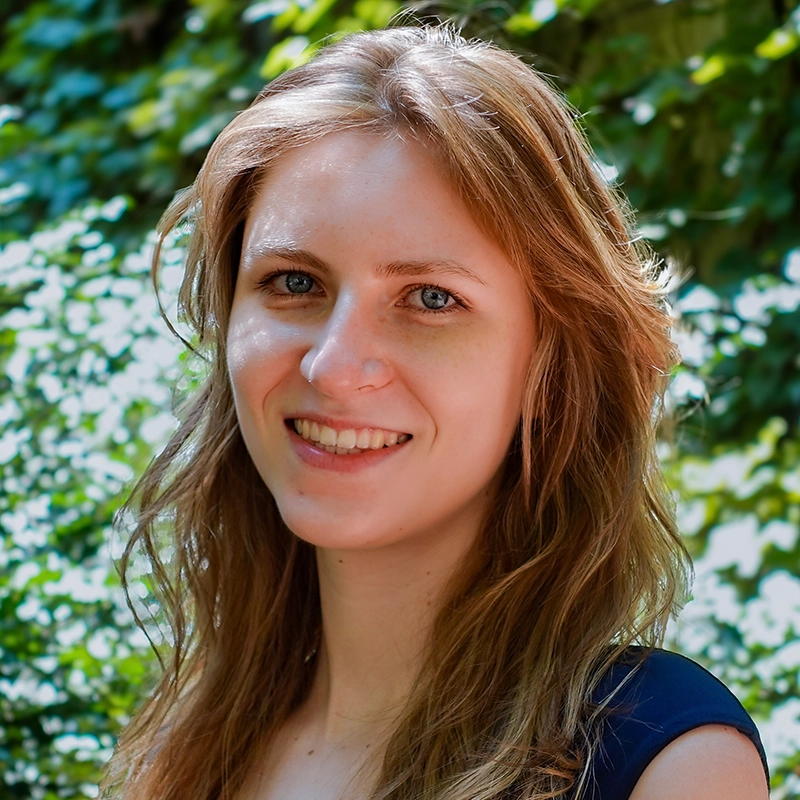 Kiana Mottahedan, a young woman with long, wavy light brown hair and fair skin, smiles at the camera. She is outdoors among lush green foliage, sunlight filtering through the leaves to create a bright, natural background.